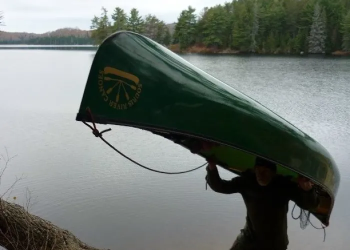 A person portaging with a canoe over the head and shoulder next to a lake